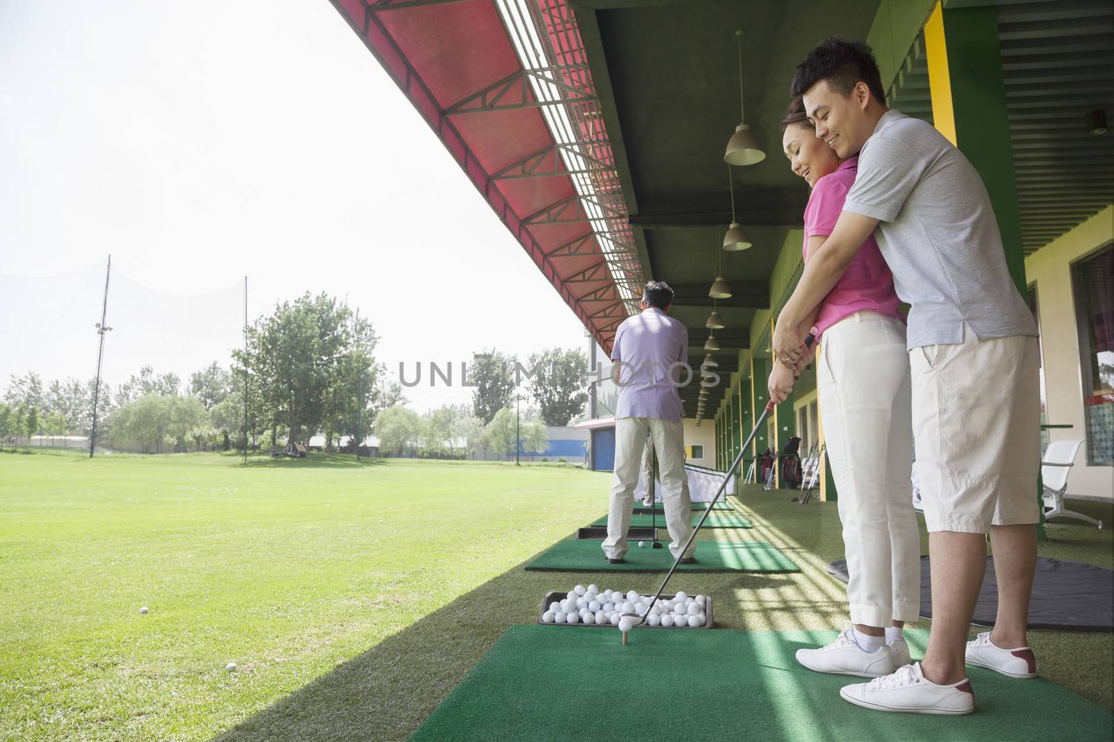 Young man teaching his girlfriend how to hit golf balls, arm around, side view