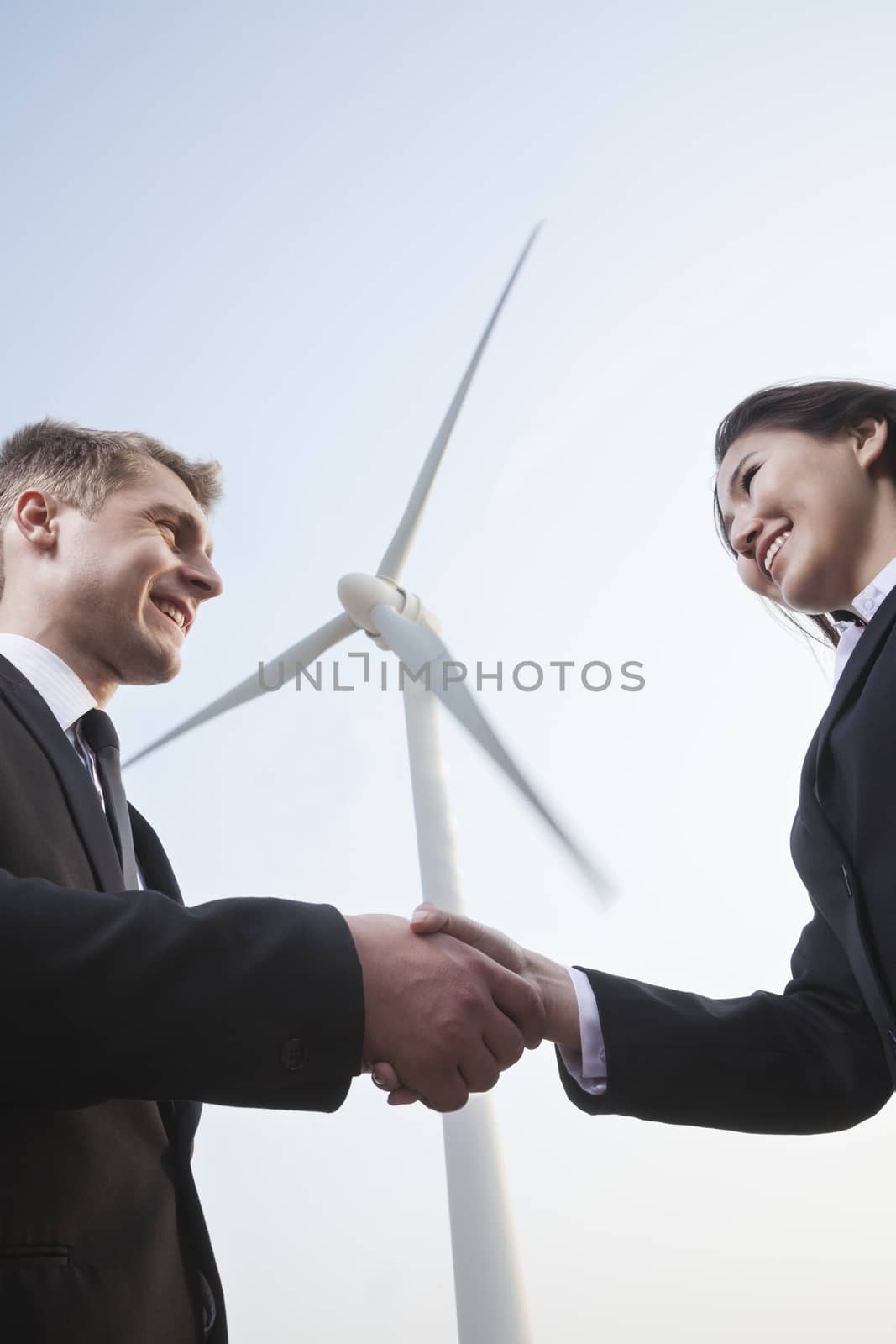 Two smiling young business people shaking hands in front of a wind turbine