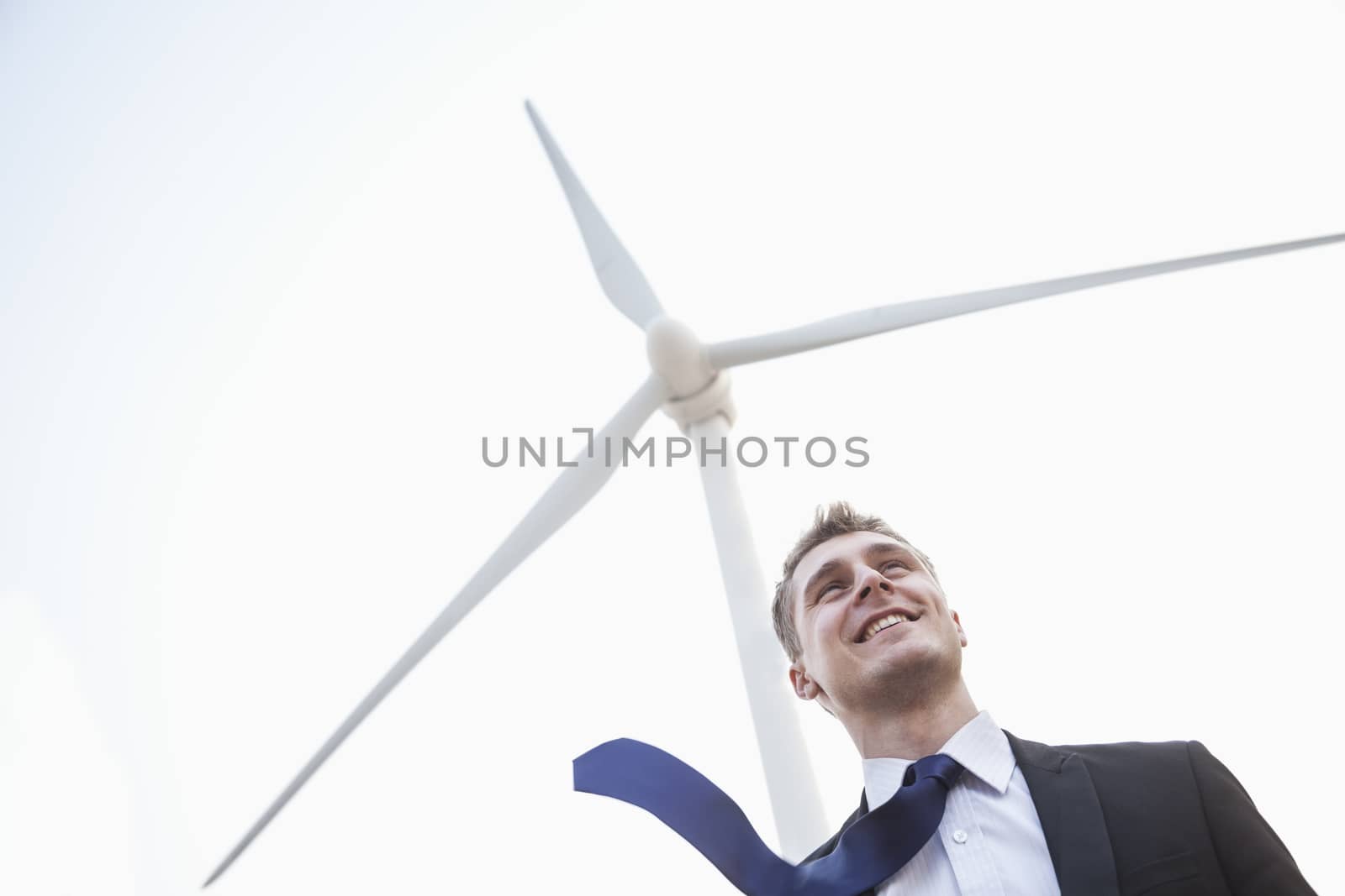 Young smiling businessman standing beside a wind turbine, tie is blowing in the wind by XiXinXing