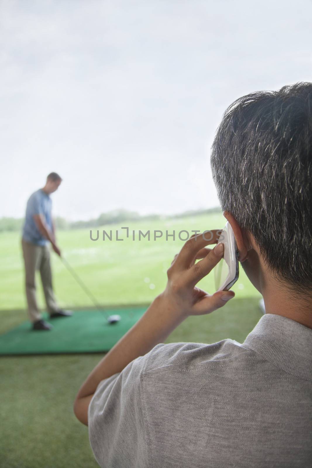Rear view of man on the phone while another man plays golf in the background by XiXinXing