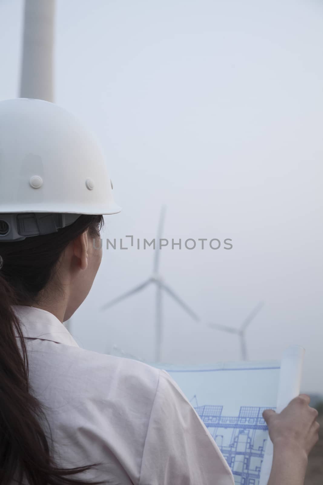 Young female engineer looking at blueprint with wind turbines in front of her, over the shoulder view by XiXinXing