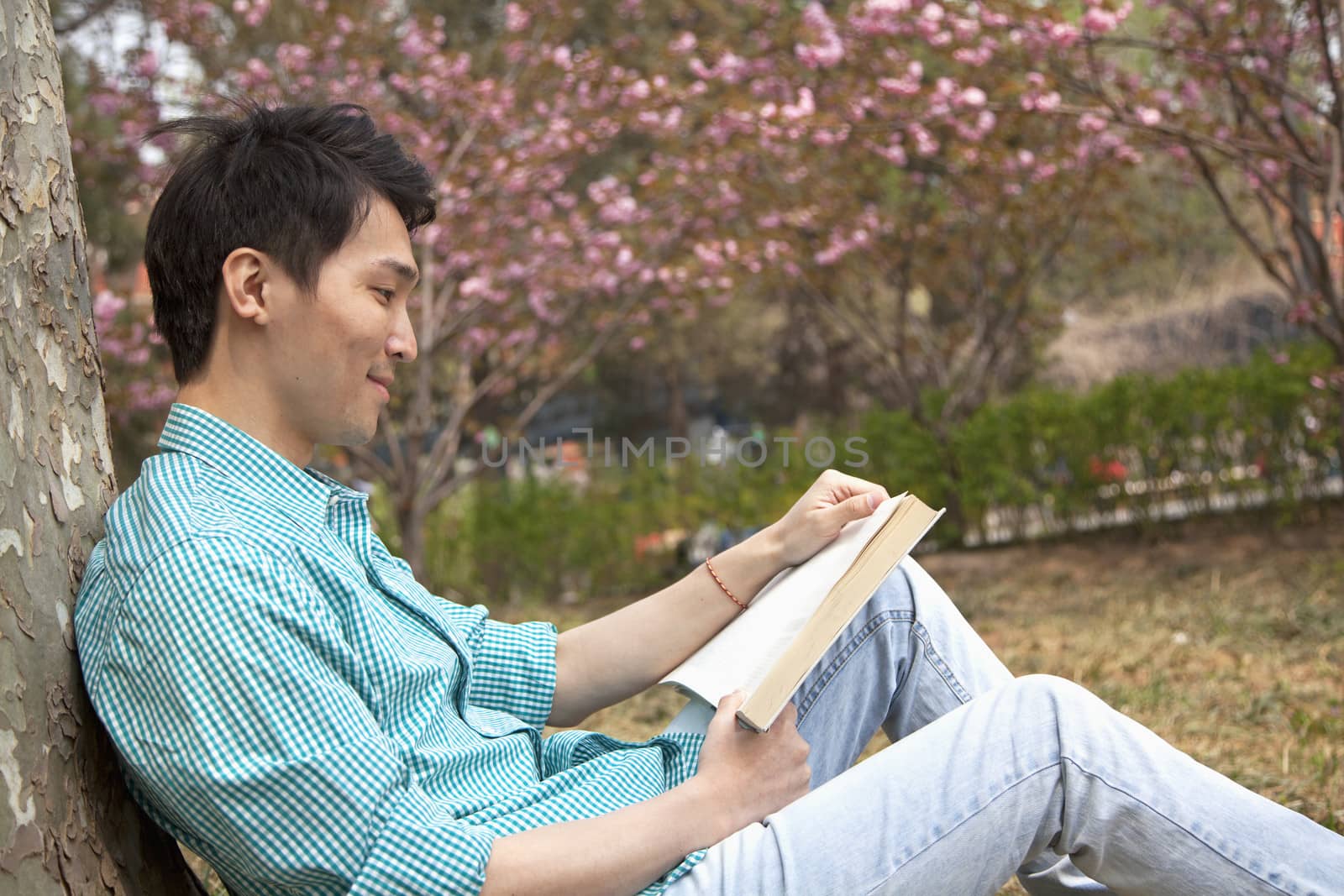 Smiling young man leaning on a tree and enjoying his book, outdoors in a park by XiXinXing