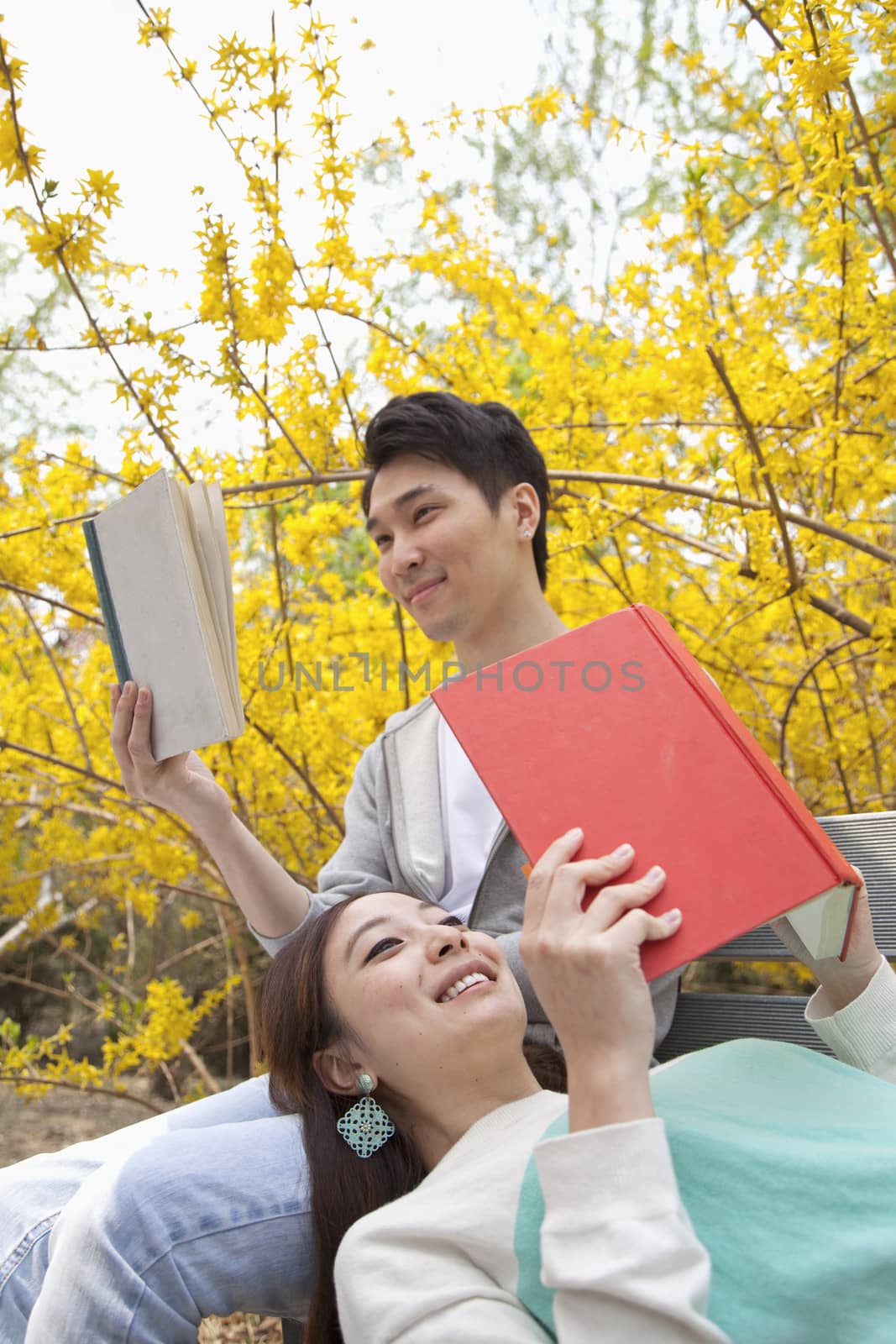 Young happy couple lying and sitting on a park bench enjoying reading their books, outdoors in springtime by XiXinXing