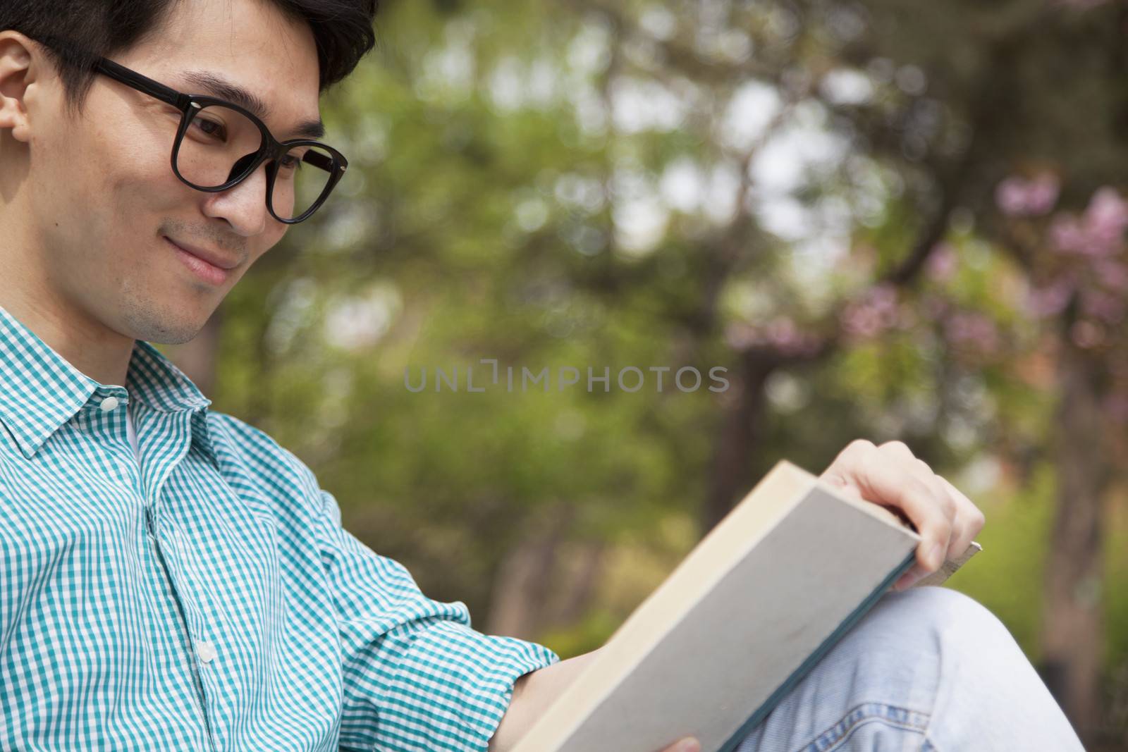 Young man with glasses smiling and enjoying his book, outdoors in a park by XiXinXing