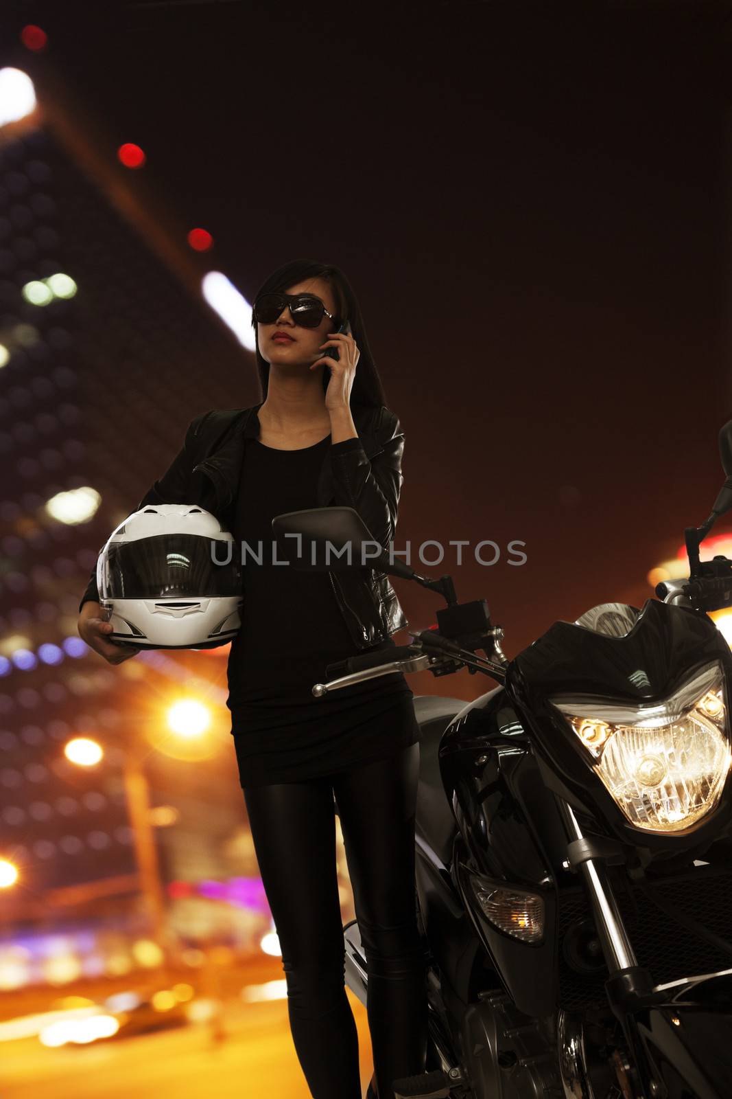 Young woman in sunglasses talking on the phone and standing beside her motorcycle at night in Beijing