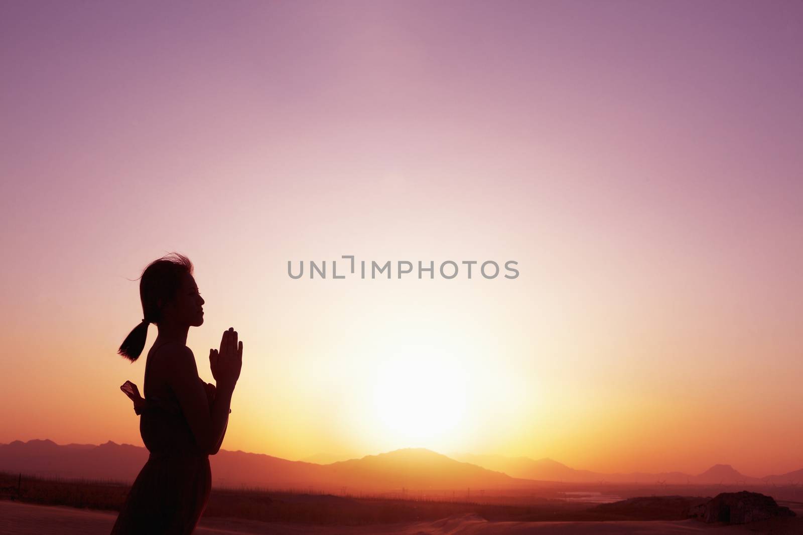 Serene young woman with hands together in prayer pose  in the desert in China, silhouette, profile, sun setting by XiXinXing