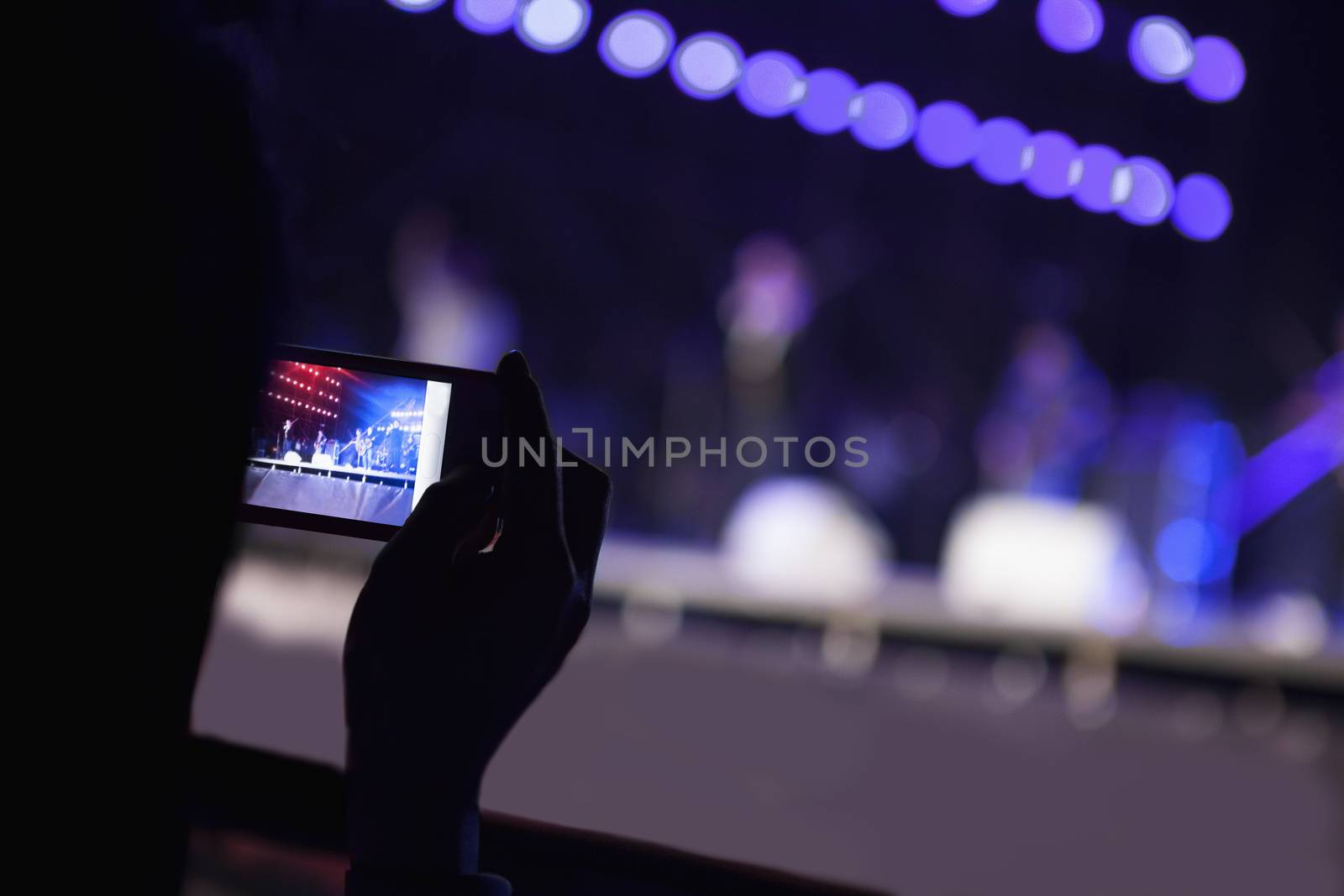 Young woman taking a photography with her smart phone at an indoor concert, over the shoulder view