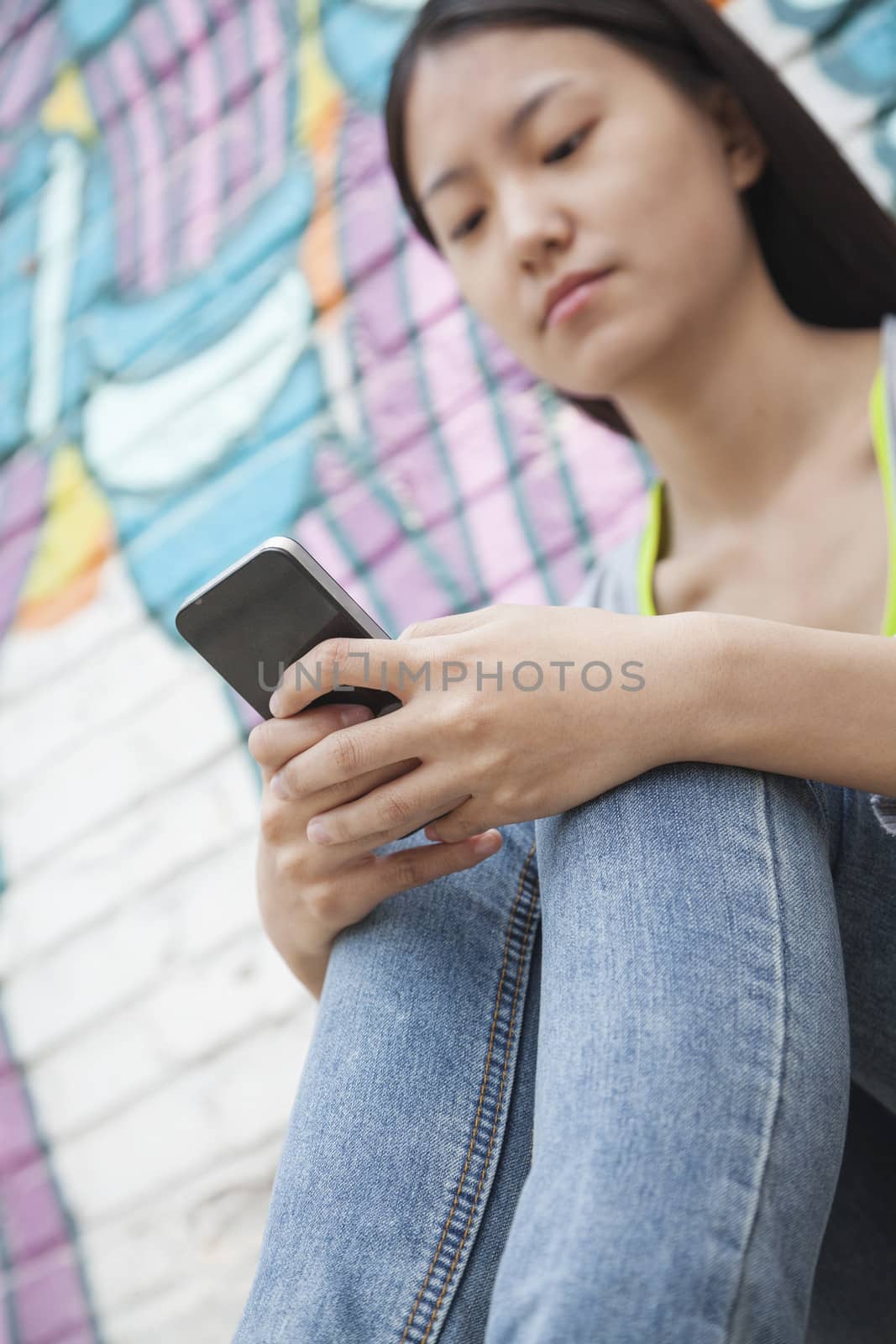Young smiling woman sitting against a wall with graffiti and looking down and texting on her phone by XiXinXing