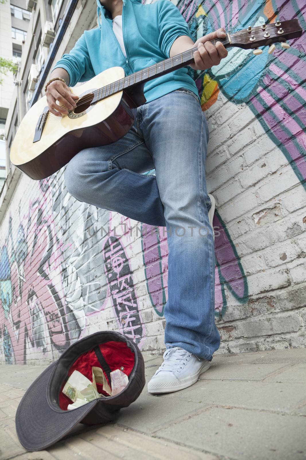 Young street musician playing guitar and busking for money in front of a wall with graffiti