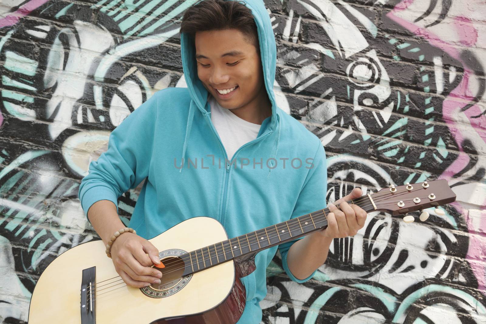 Young smiling street musician leaning on a wall with graffiti drawings and playing his guitar