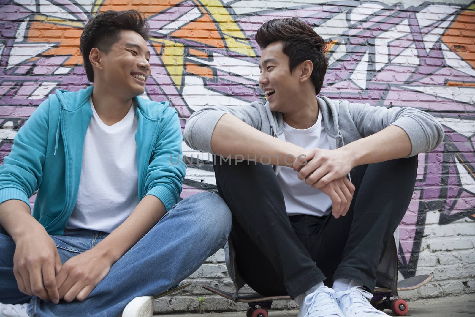 Two young men sitting on their skateboards and hanging out in front of a wall with graffiti by XiXinXing