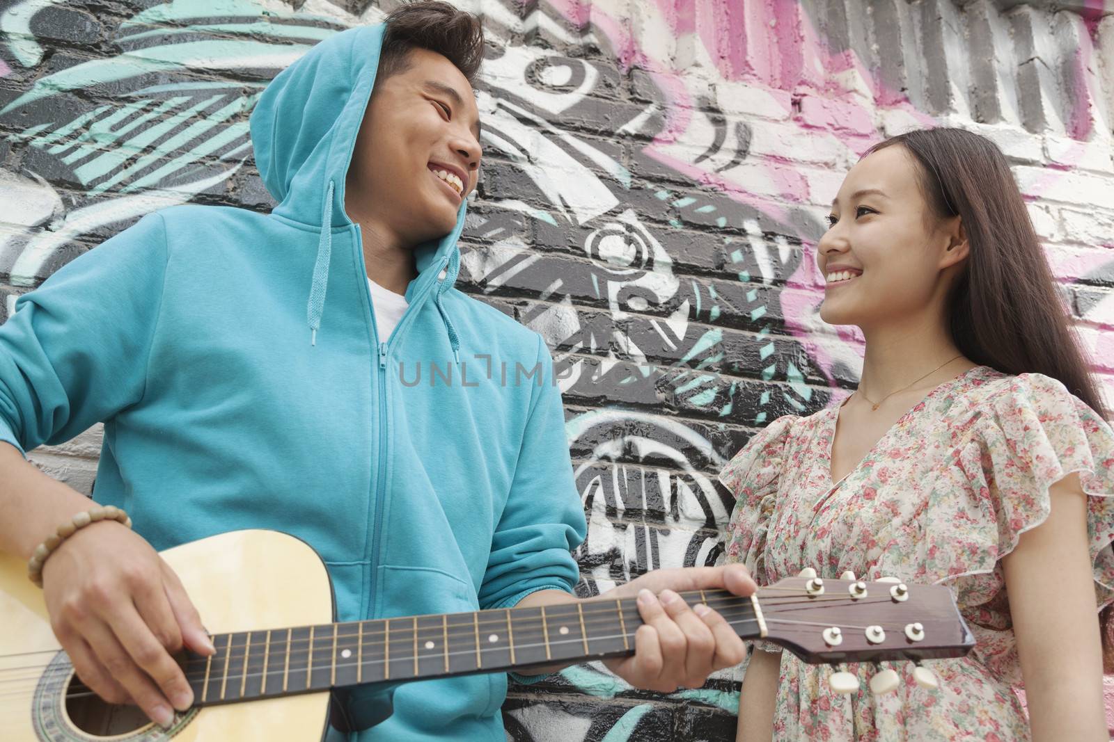 Young smiling street musician leaning on a wall with graffiti drawings, playing his guitar, and flirting with a young woman in a dress