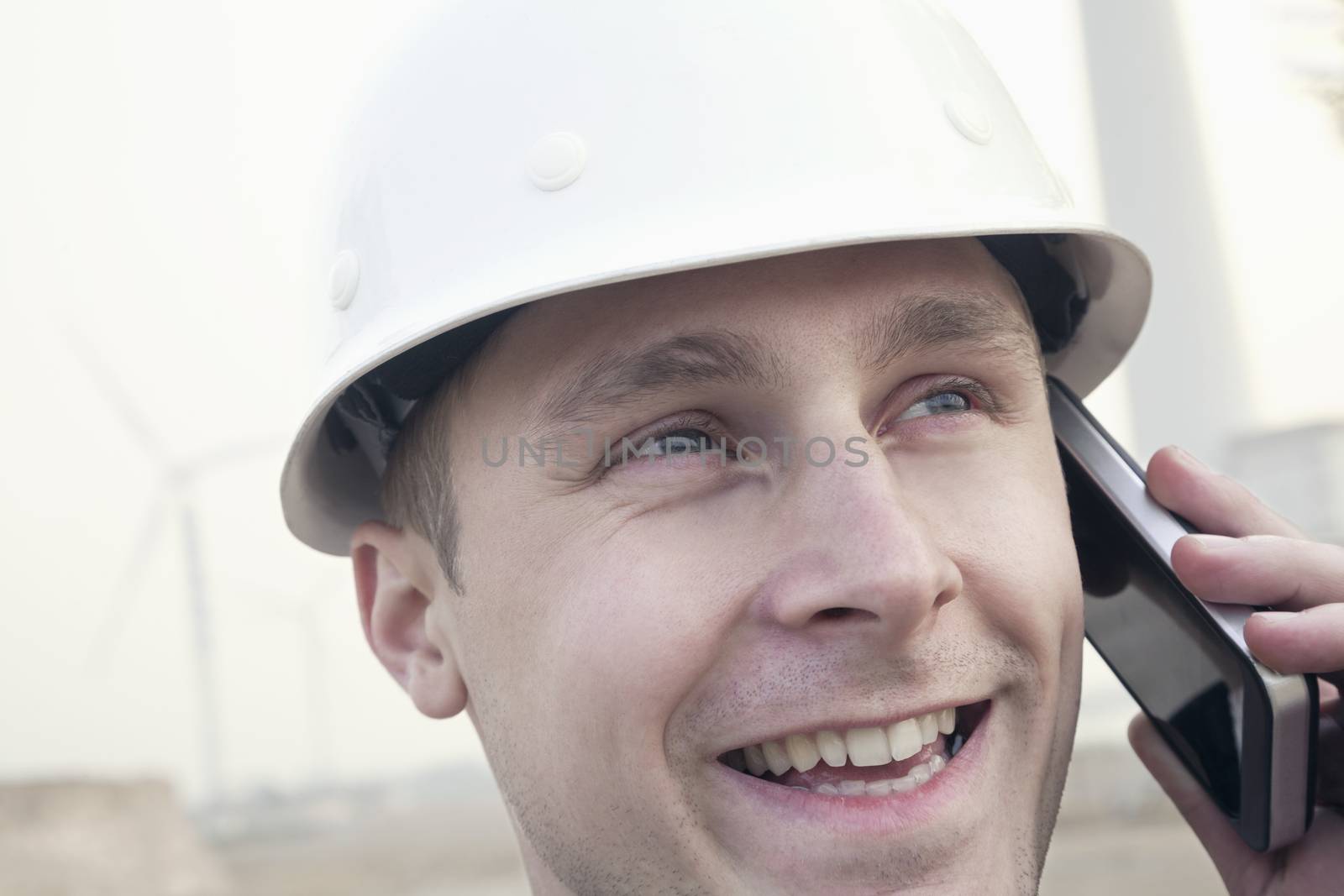 Young smiling male engineer in a hardhat on the phone beside a wind turbine, close-up by XiXinXing
