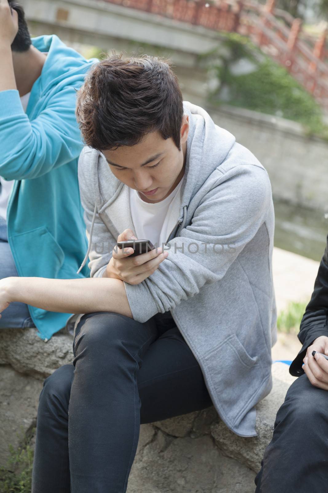 Young man in a gray hooded sweatshirt looking down at his phone and texting outdoors by XiXinXing