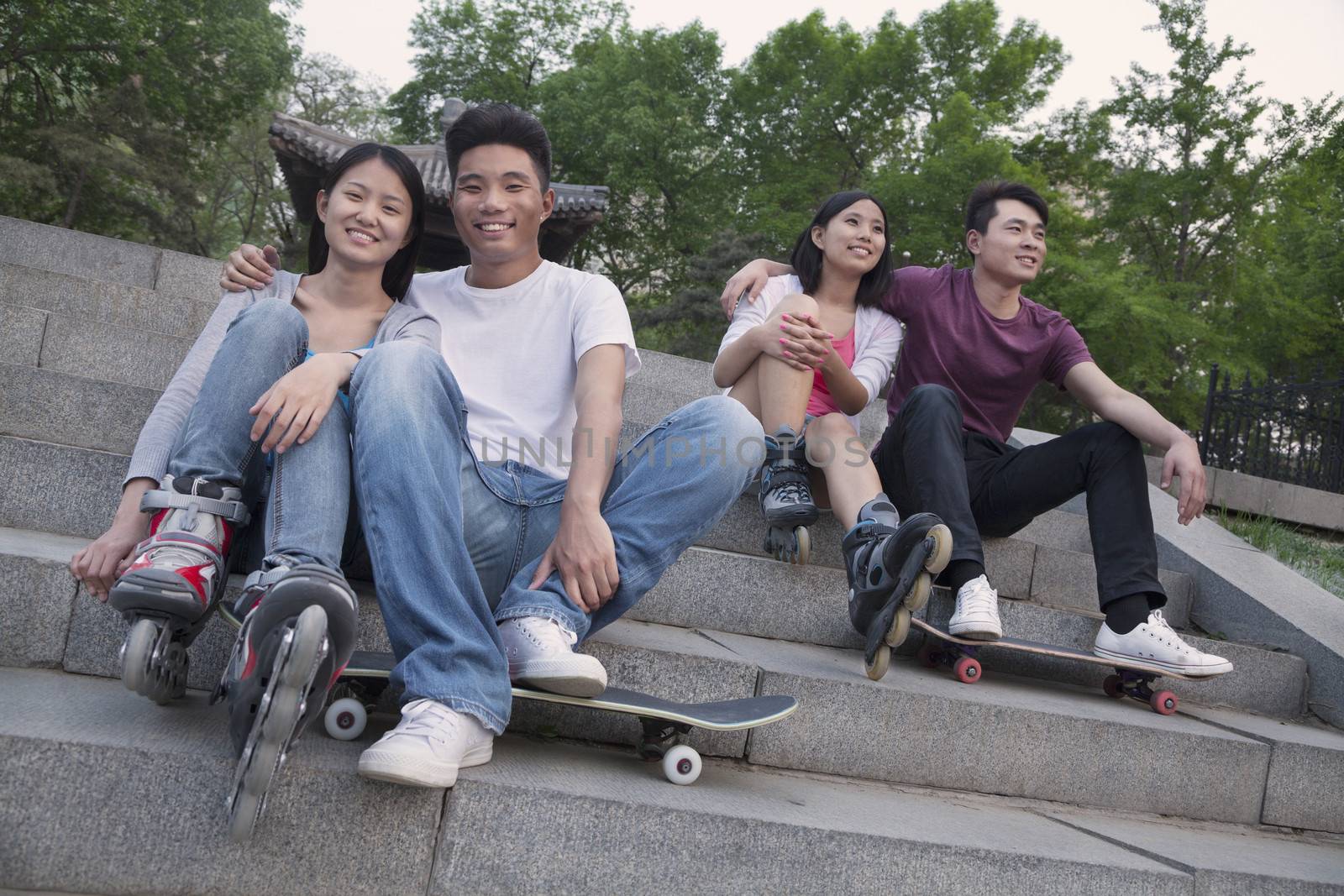 Two young couples sitting and resting on concrete steps outside with skateboards and roller blades by XiXinXing