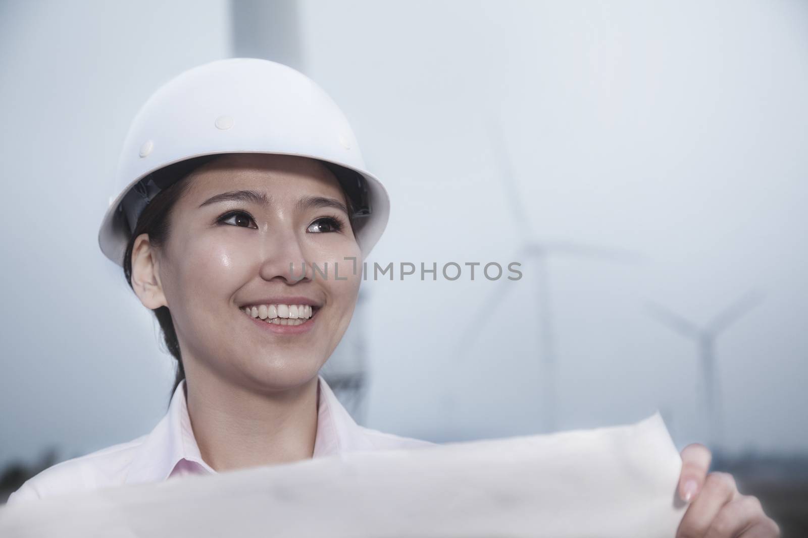 Young smiling female engineer holding a blueprint with wind turbines in the background by XiXinXing