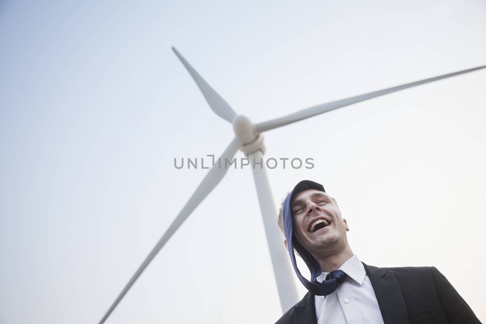 Young smiling businessman standing beside a wind turbine, tie is on his head