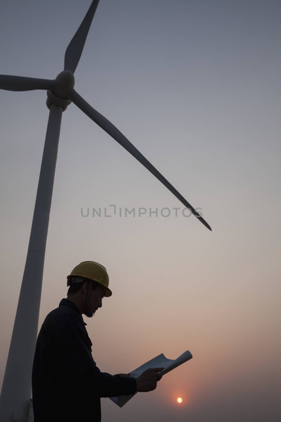 Silhouette of young male engineer holding a blueprint and standing beside a wind turbine at sunset by XiXinXing