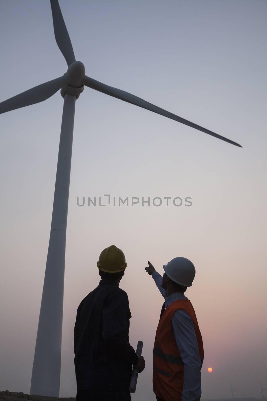 Two young male engineers standing beside a wind turbine at sunset, pointing up  by XiXinXing