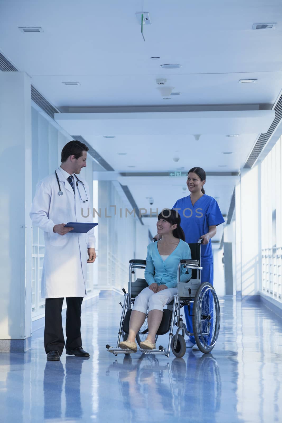 Smiling female nurse pushing and assisting patient in a wheelchair in the hospital, talking to doctor by XiXinXing