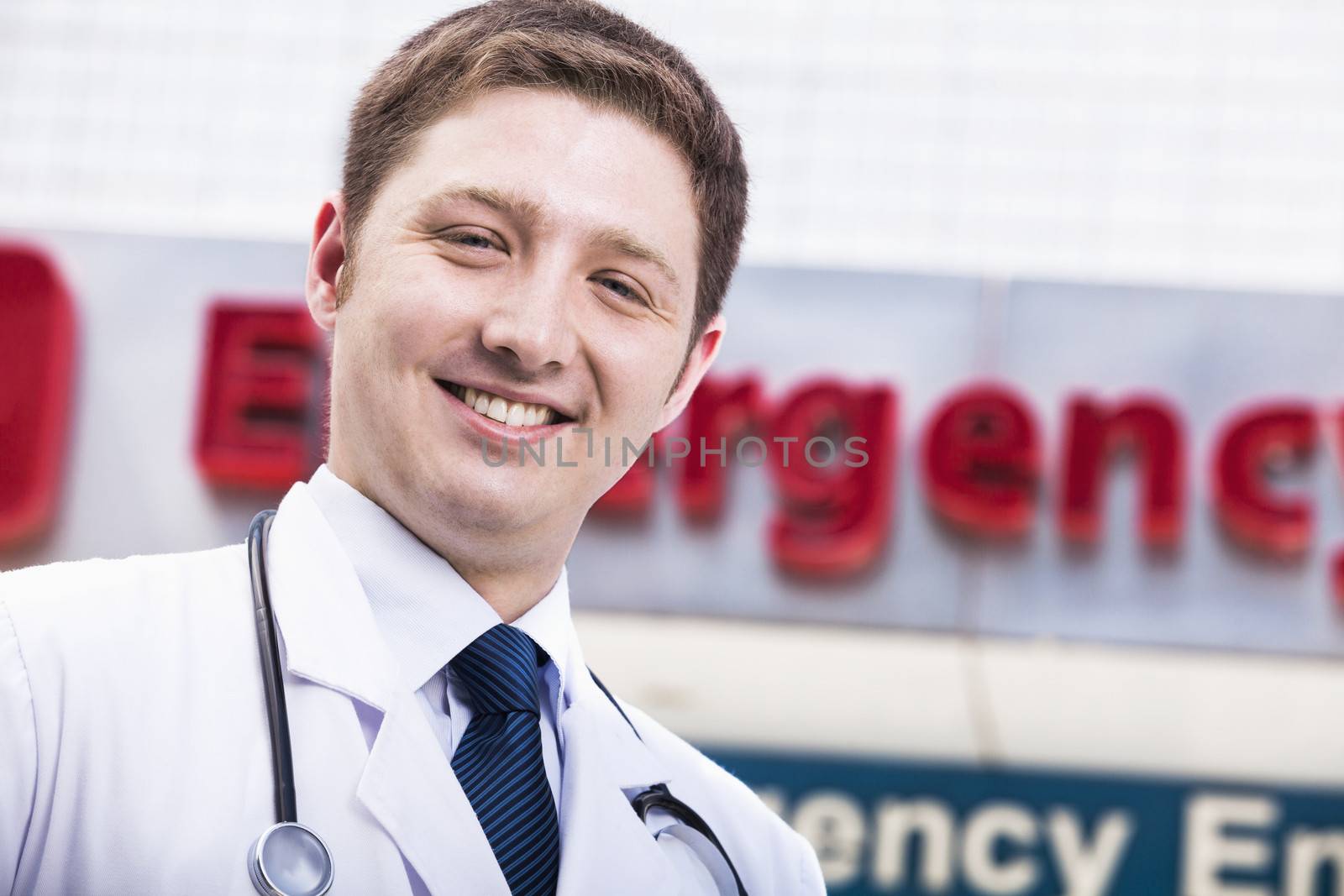 Portrait of young smiling doctor outside of the hospital, emergency room sign in the background by XiXinXing