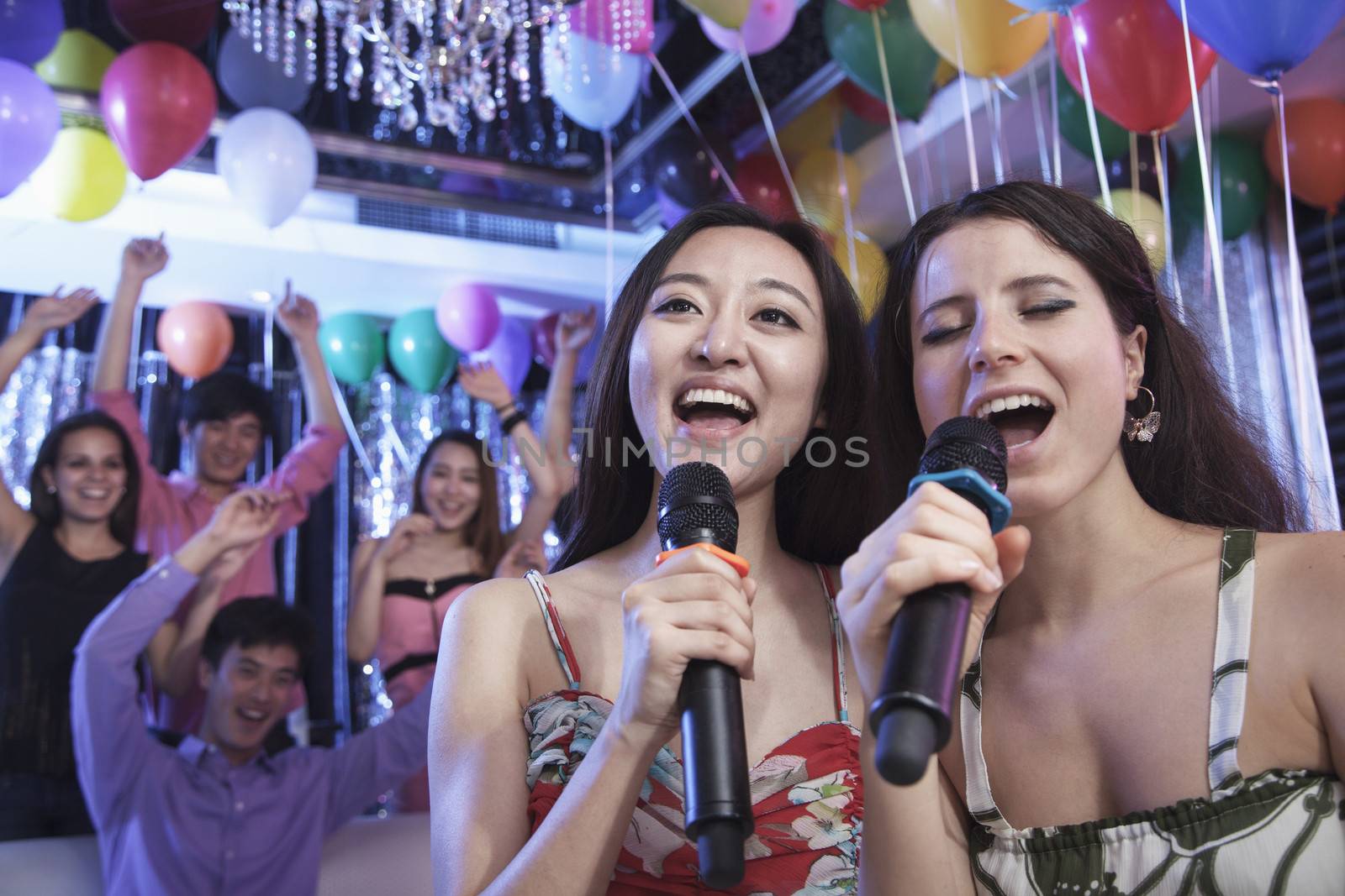 Two friends holding microphones and singing together at karaoke, friends in the background