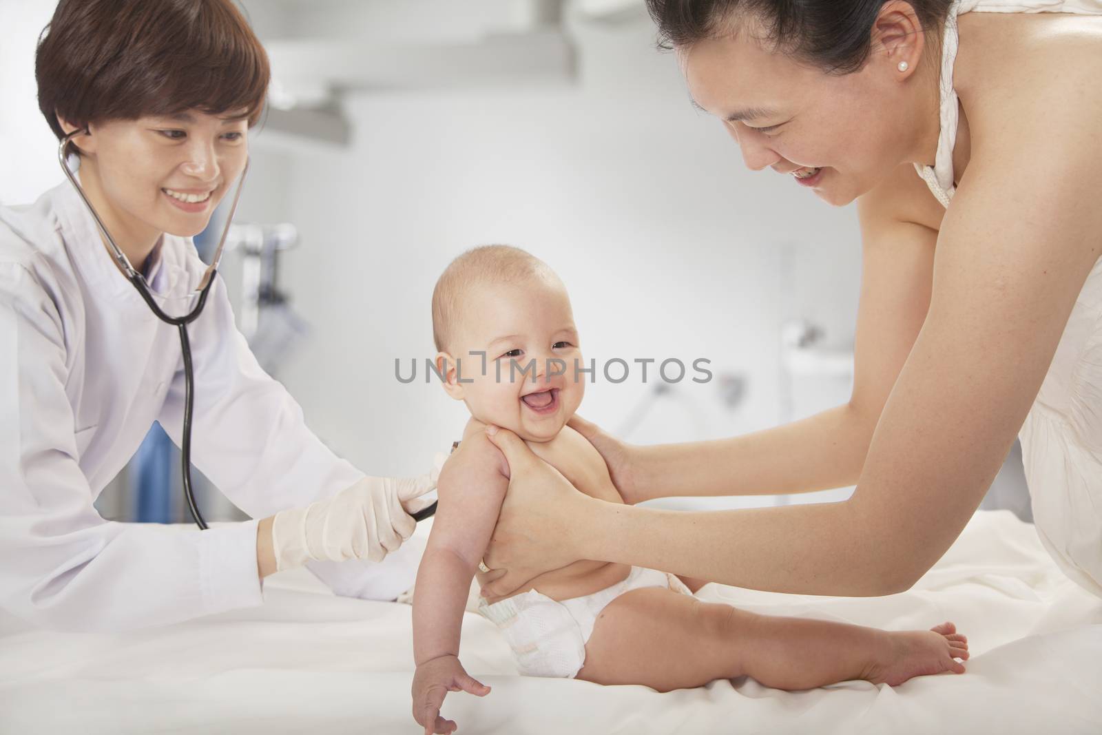Doctor examining the baby with a stethoscope in the doctors office, mother holding the baby by XiXinXing