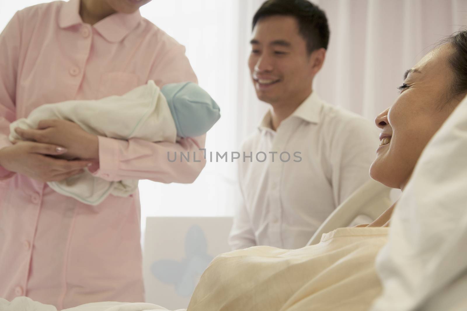 Nurse holding newborn baby in the hospital with mother lying on the bed and the father beside her