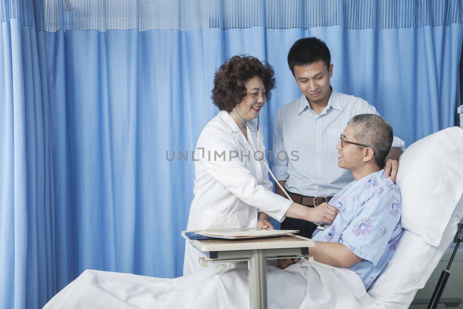 Doctor checking up on patient who is lying down in bed with adult son by his side