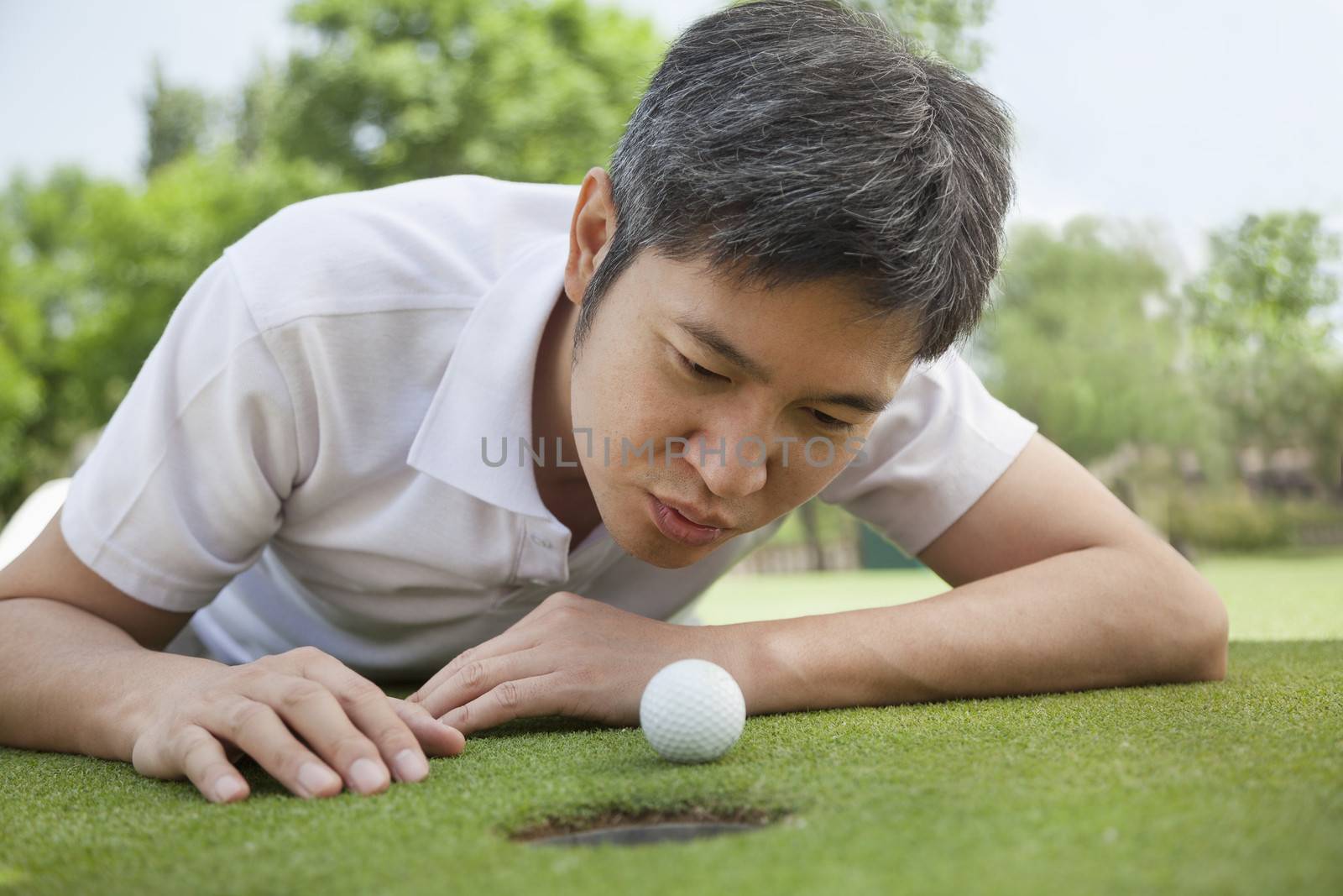 Mid adult man lying down in a golf course trying to blow the ball into the hole