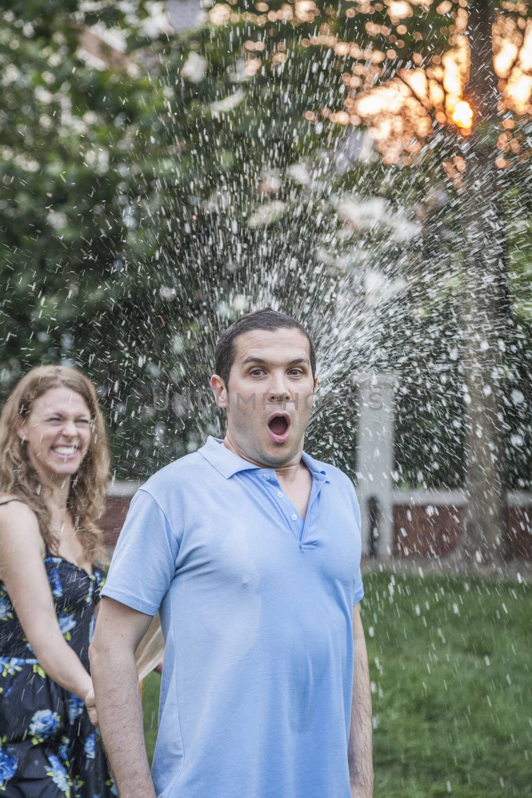 Couple playing with a garden hose and spraying each other outside in the garden, man has a shocked look 