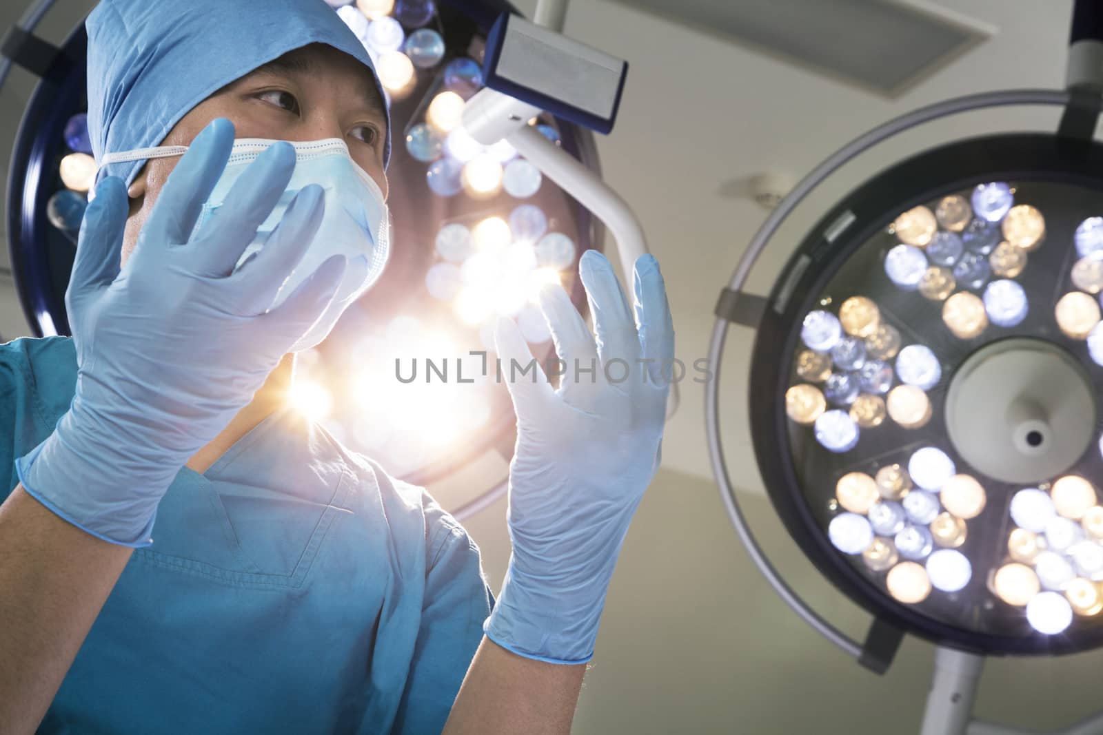 Low angle view of surgeon holding gloved hands up with surgical lights behind him by XiXinXing