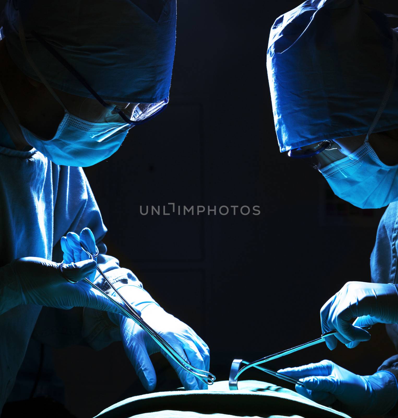 Two surgeons looking down, working, and holding surgical equipment with patient lying on the operating table 