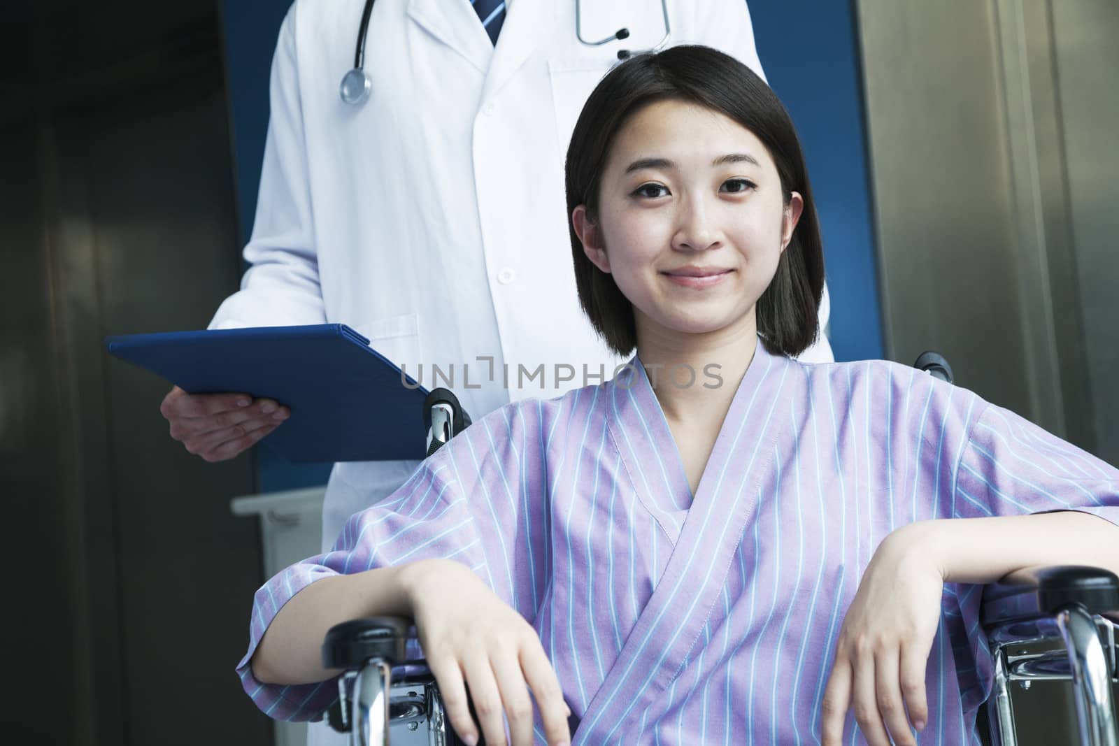 Young smiling female patient sitting in a wheelchair, doctor standing behind her, looking at camera