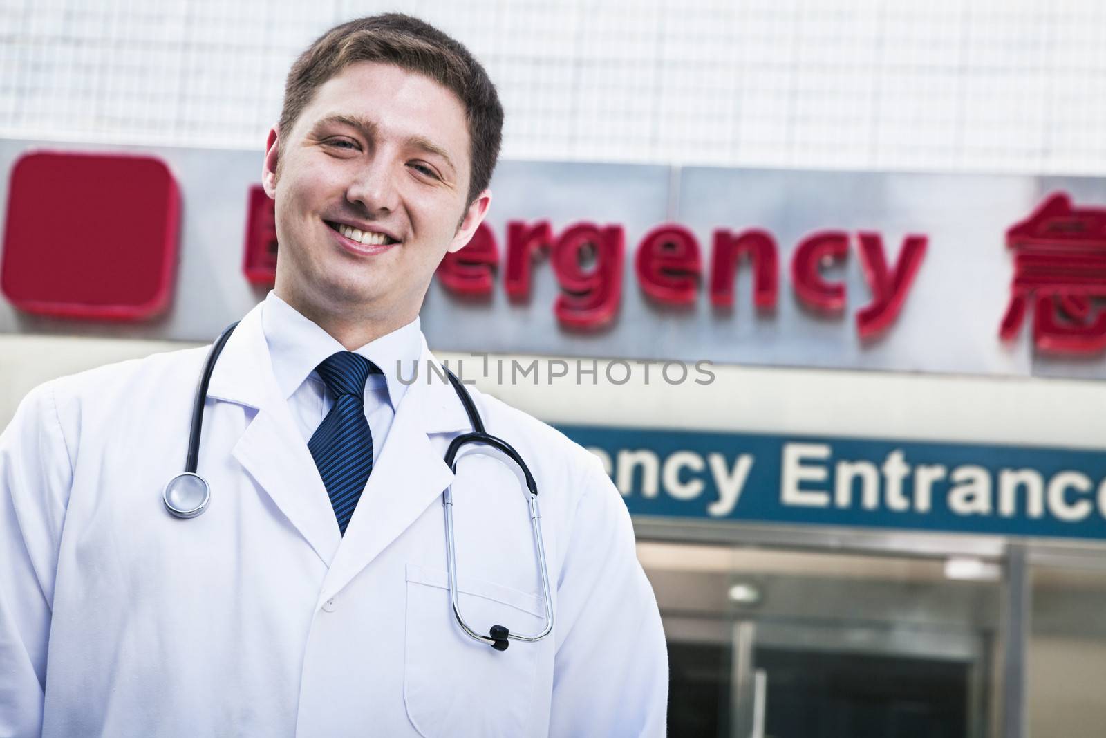 Portrait of young smiling doctor outside of the hospital, emergency room sign in the background