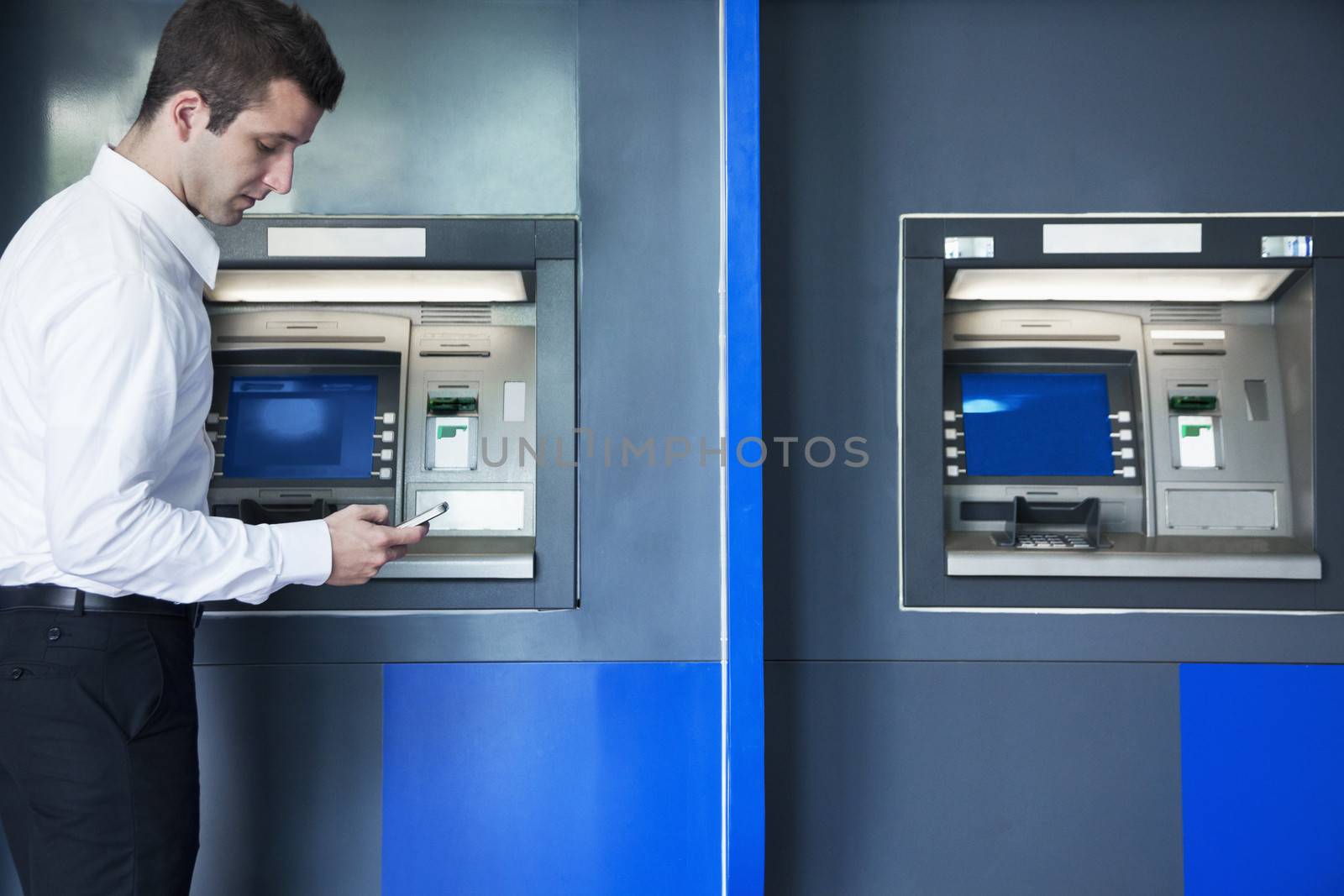 Young businessman taking out money from the ATM and looking down at his phone