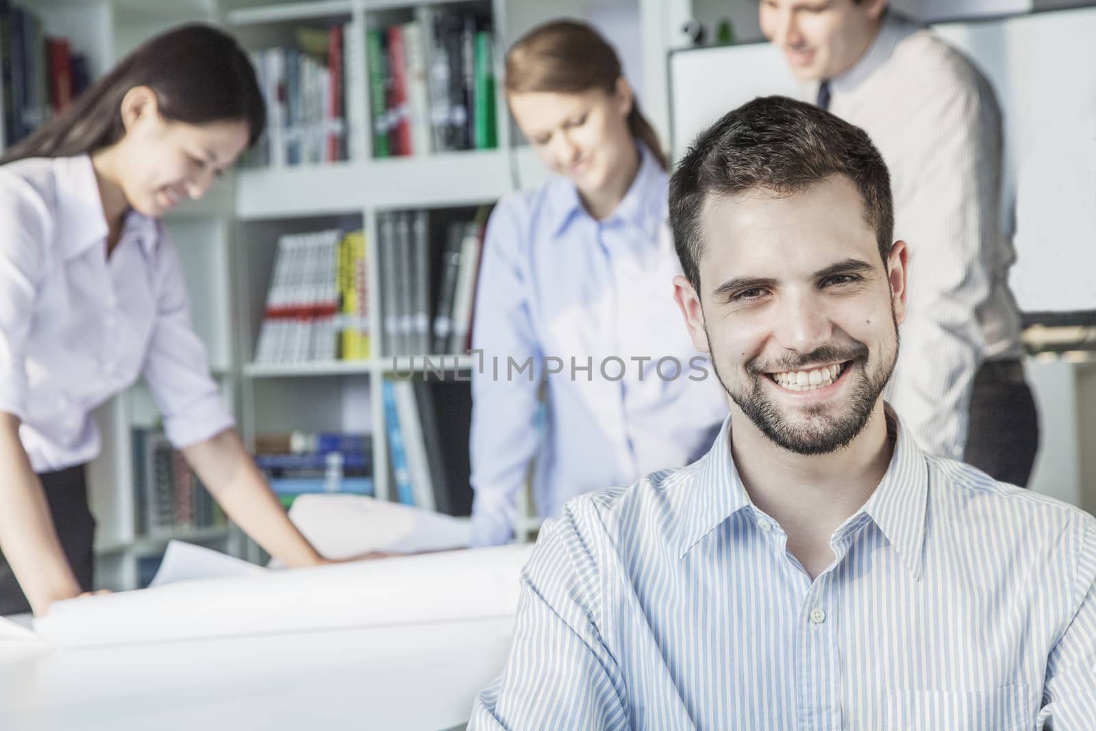Smiling architect looking at camera while colleagues are planning around a table and looking at a blueprint by XiXinXing