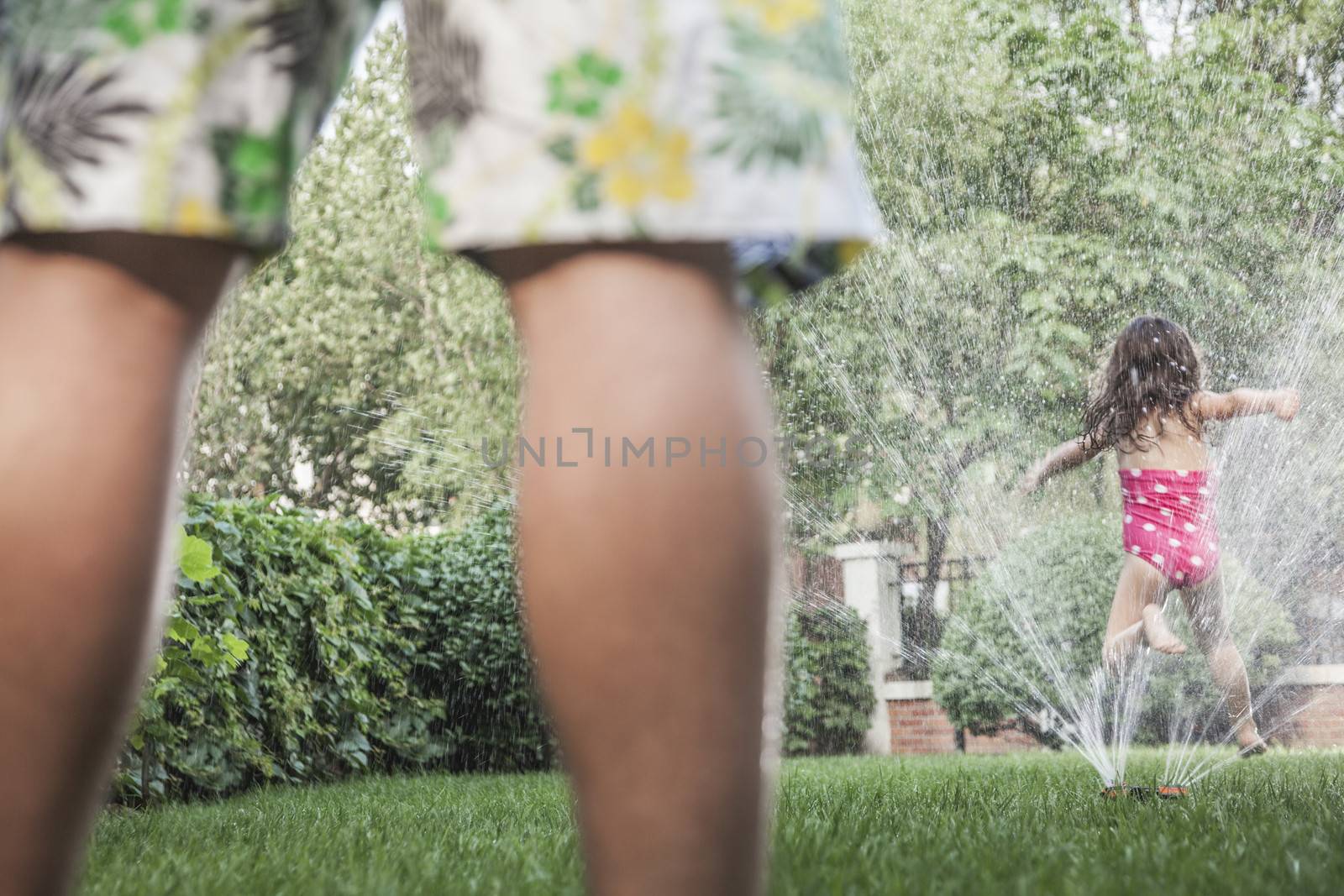 Little girl jumping through a sprinkle as father watched, low section