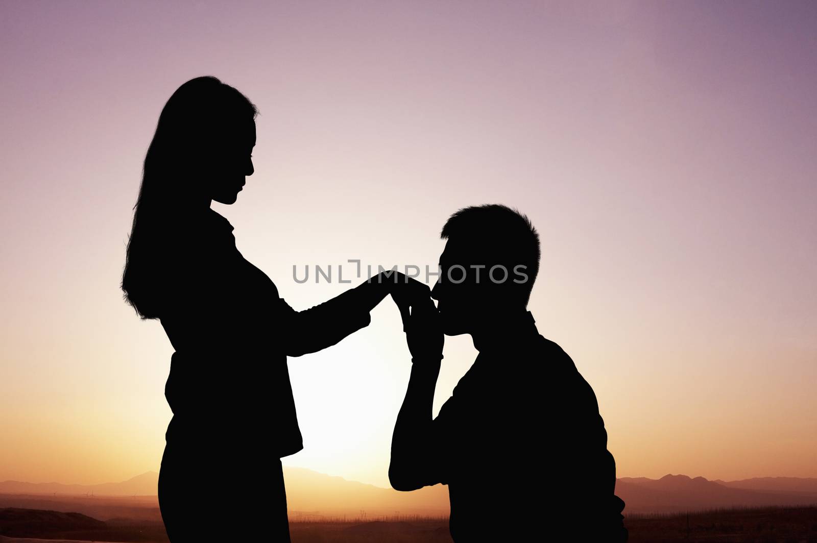 Silhouette of boyfriend kneeling and kissing his girlfriends hand at sunset