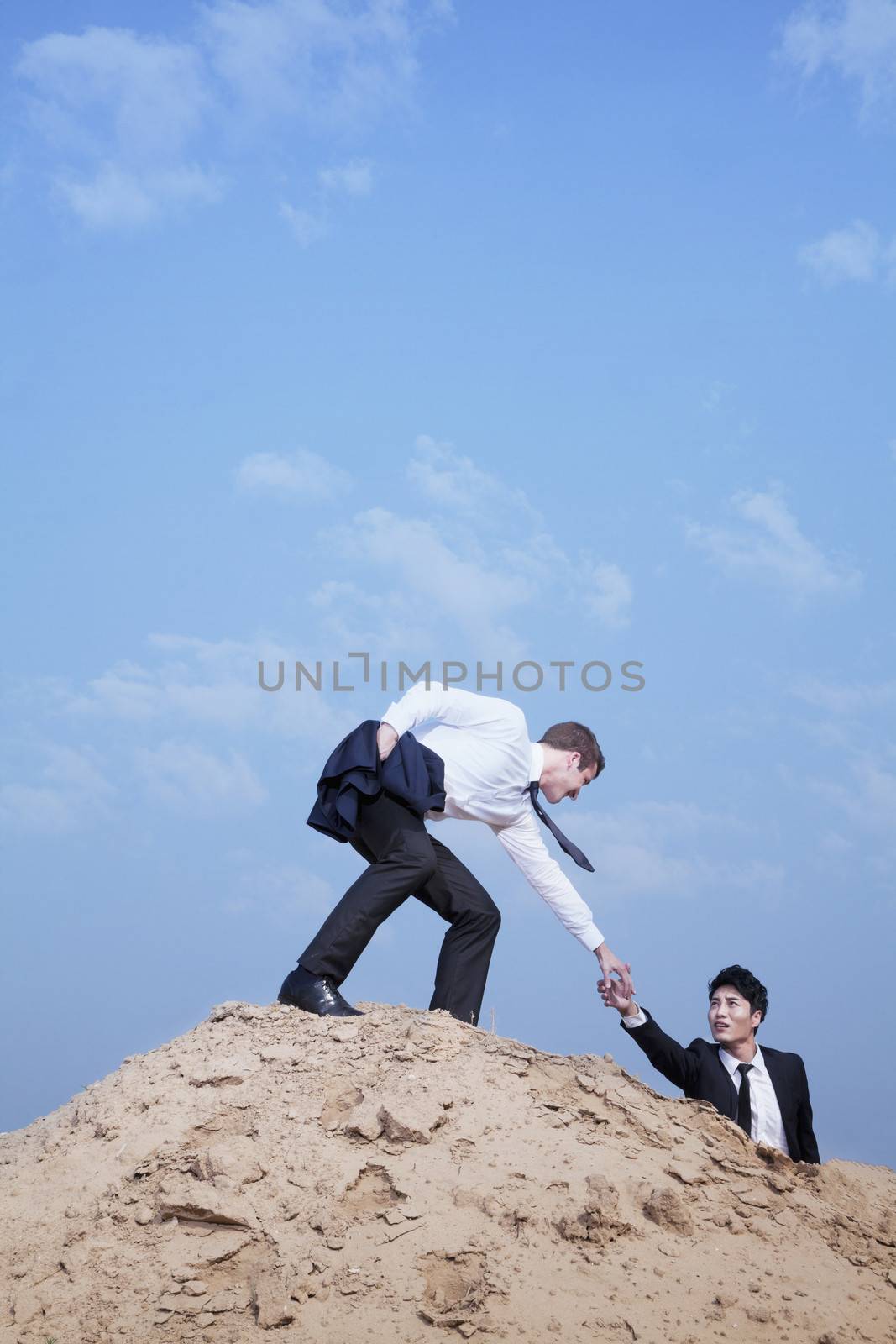 Young businessman helping another businessman get to the top of the hill in the desert by XiXinXing
