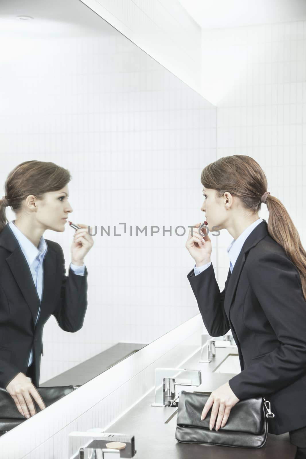 Young businesswoman with long hair looking into the mirror and putting on lipstick in the bathroom