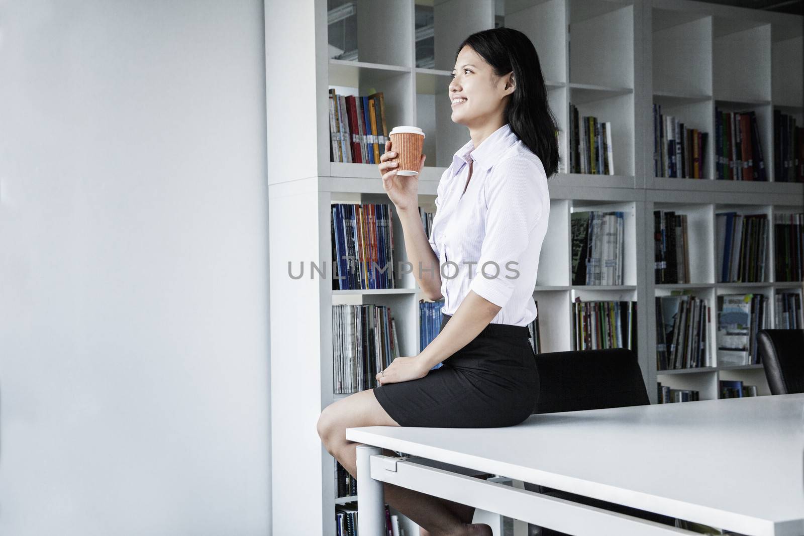 Smiling young businesswoman holding a coffee and sitting on a table, looking away