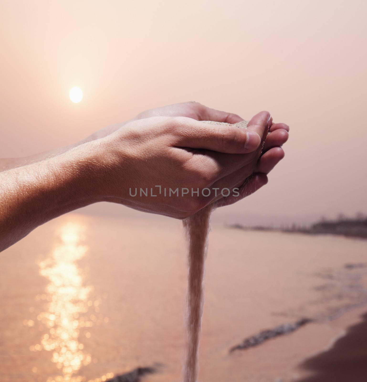 Hands holding and spilling sand with beach at sunset in the background by XiXinXing