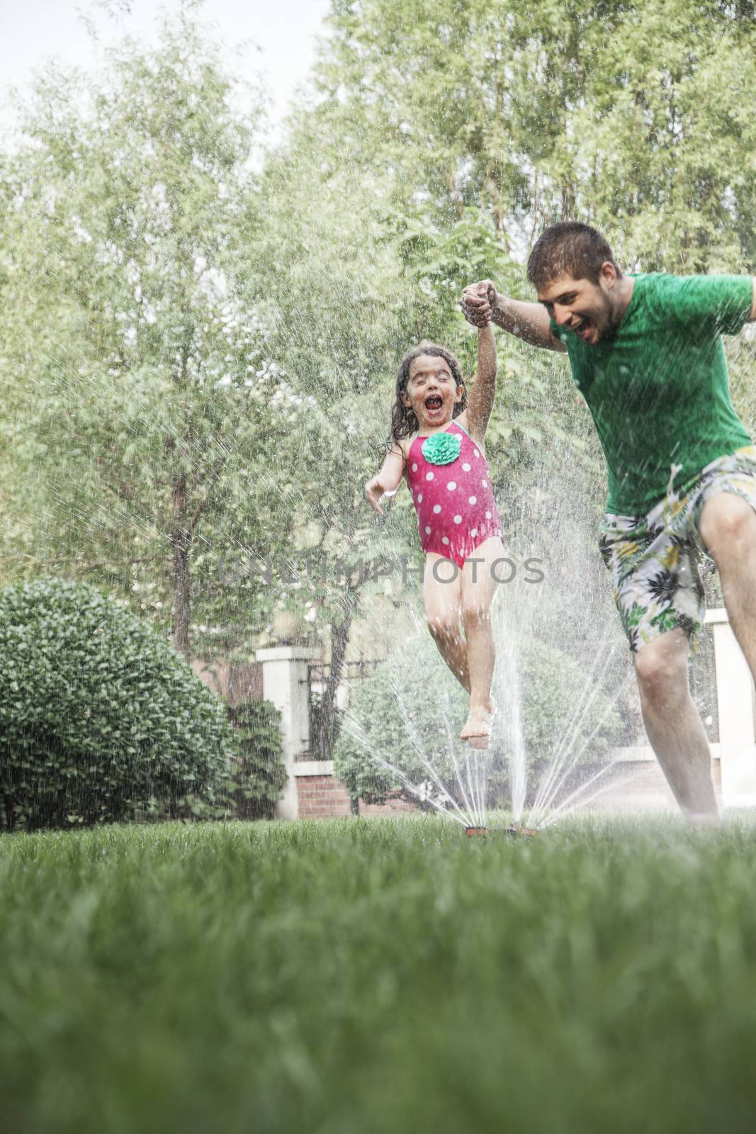 Father holding daughters hand while she jumps through the sprinkler in the garden by XiXinXing