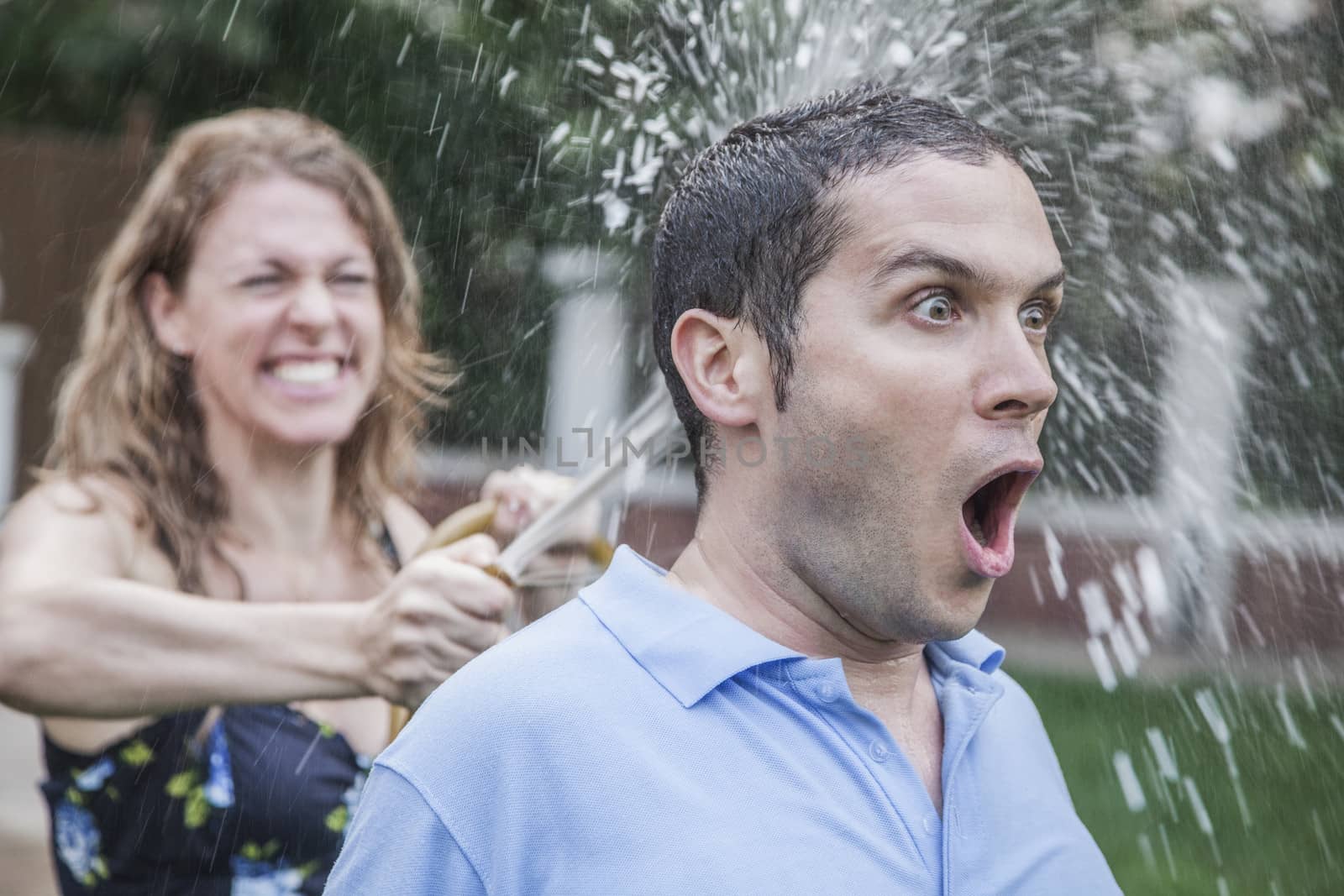 Couple playing with a garden hose and spraying each other outside in the garden, man has a shocked look, close-up 