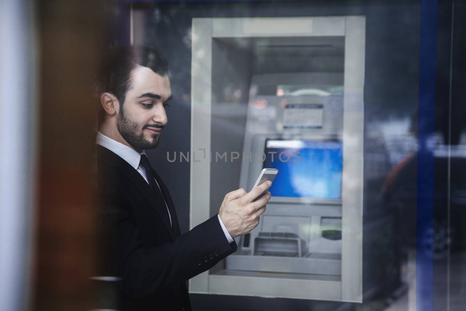 Smiling young businessman standing in front of an ATM and looking at his phone by XiXinXing