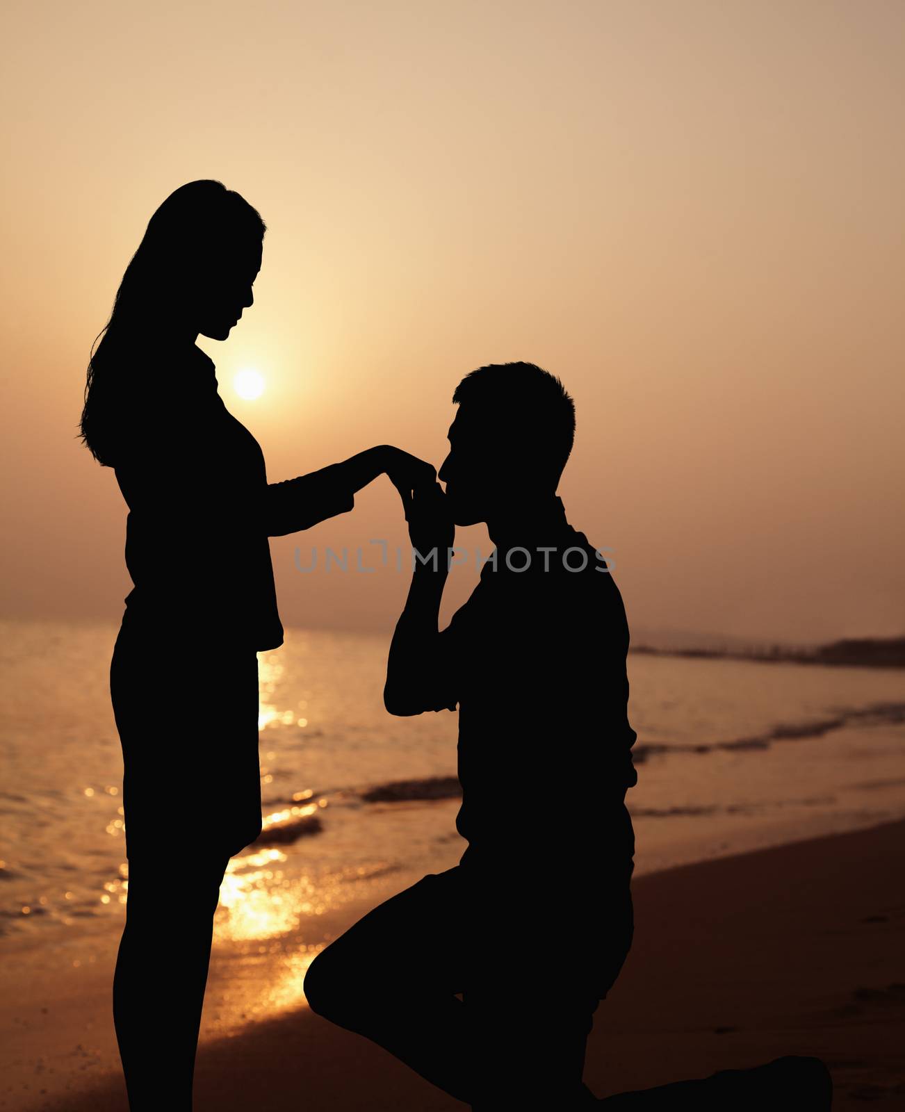 Silhouette of boyfriend kneeling and kissing his girlfriends hand on the beach at sunset