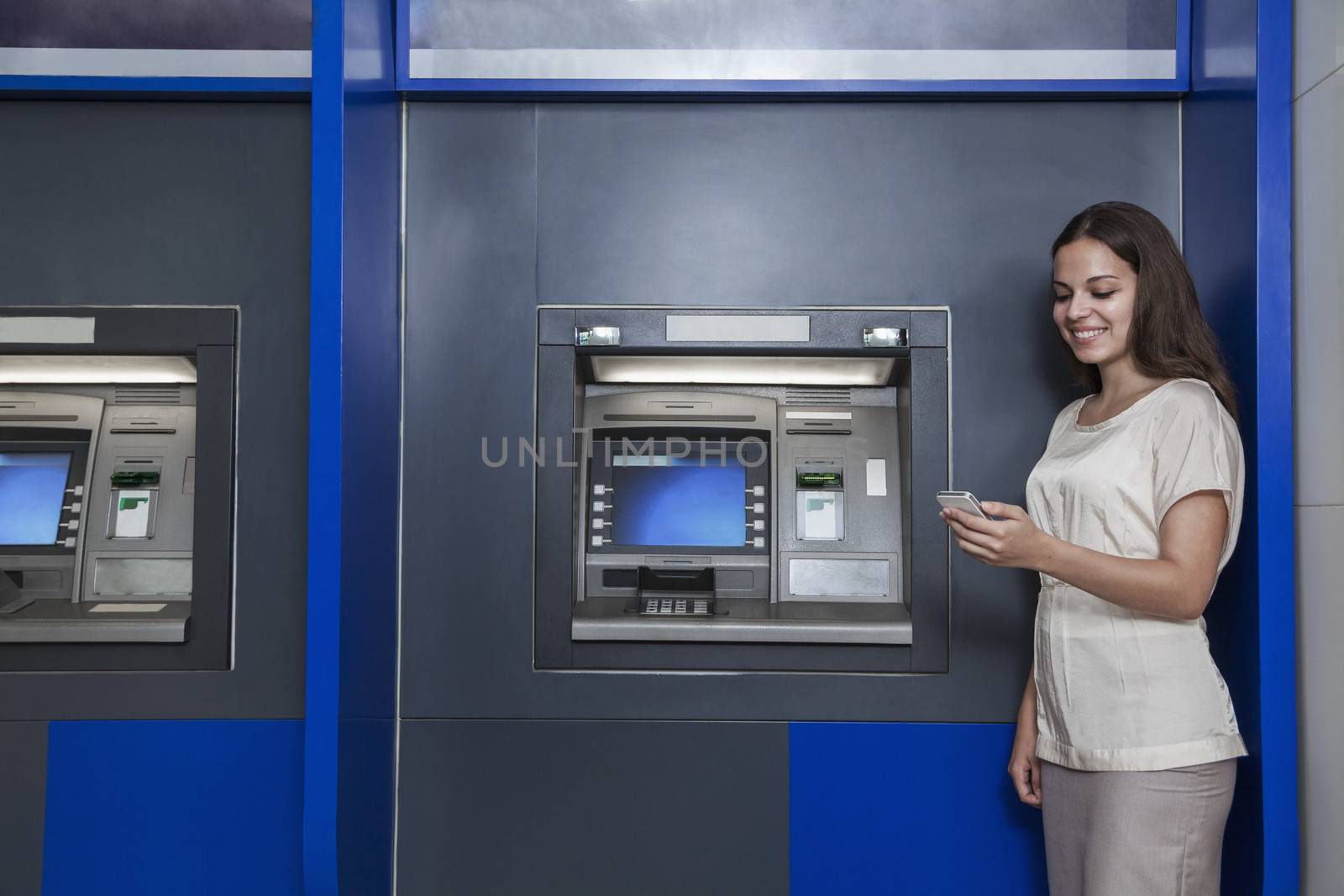 Smiling young woman standing in front of an ATM and looking at her phone by XiXinXing