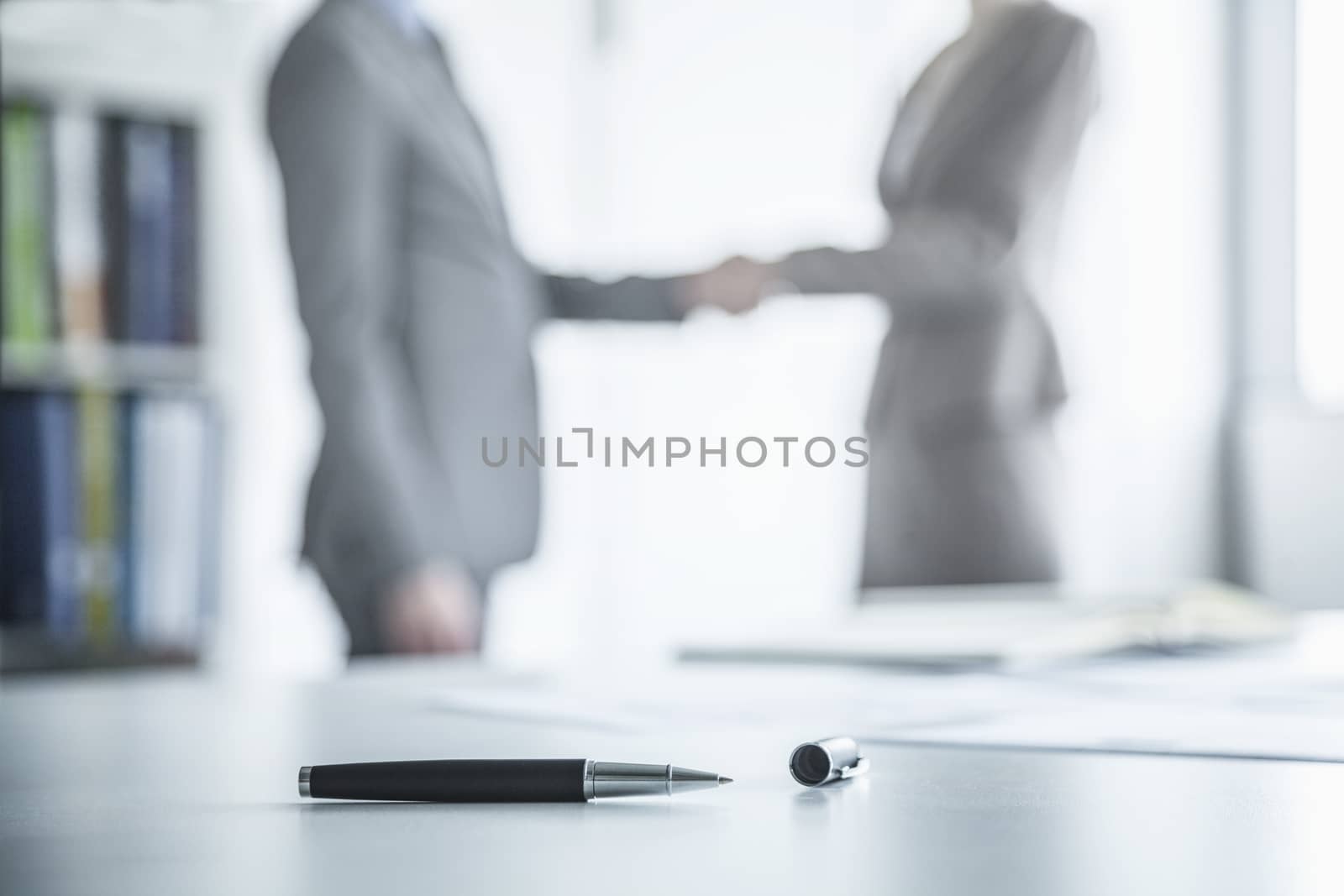 Two business people shaking hands in the background, pen lying on the table in the foreground
