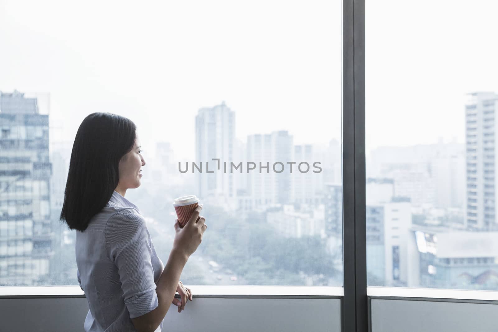 Smiling young businesswoman holding a coffee cup and looking out the window a the cityscape in Beijing, China by XiXinXing